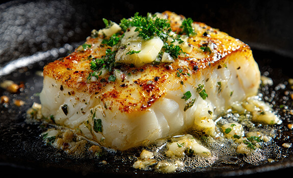 Cube of Cod grilled with pat of butter and herbs on top in iron skillet